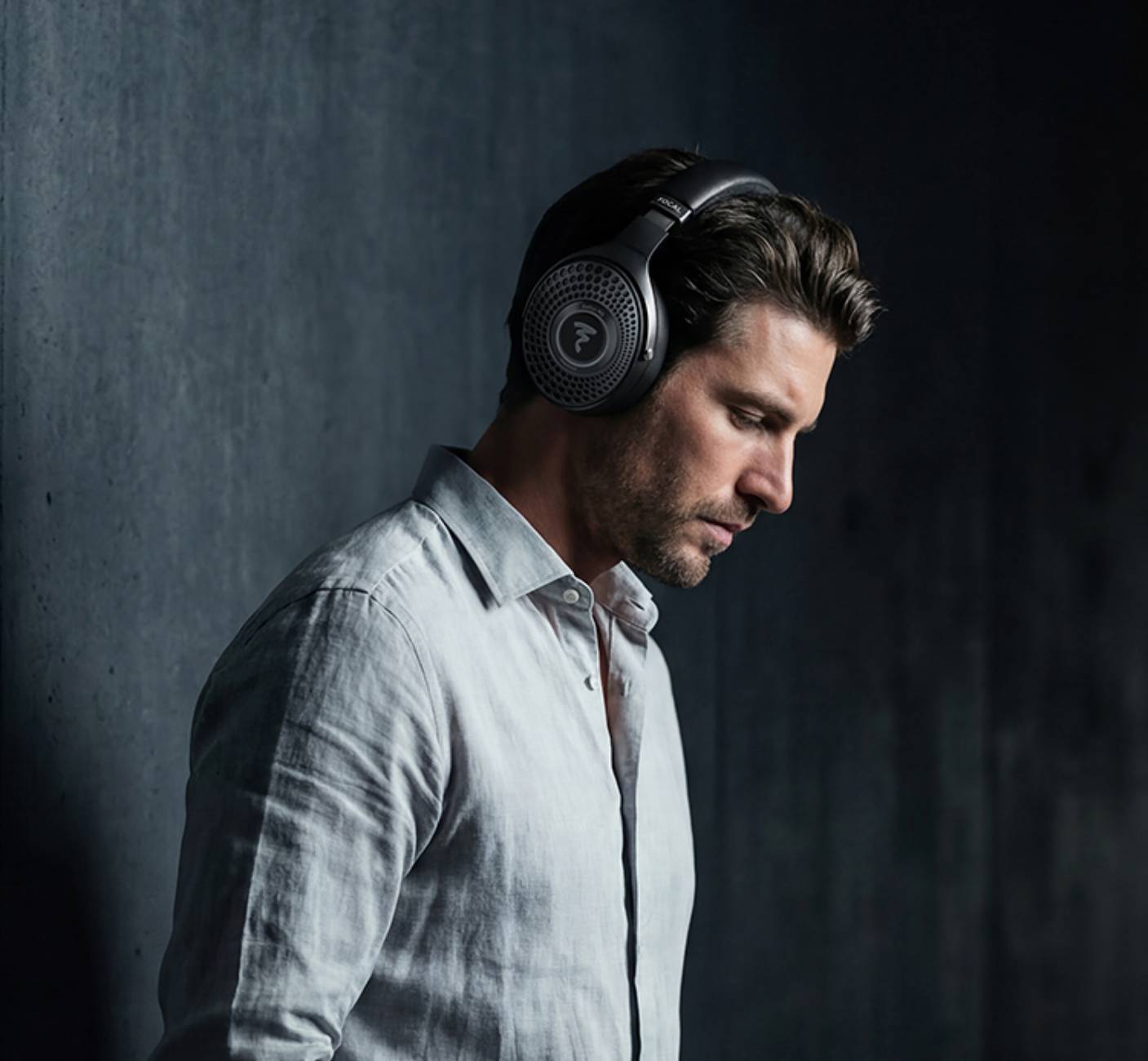 Gros plan sur l'oreillette circumaurale fermée du casque Focal Bathys MG en finition Graphen Grey, montrant la grille perforée en aluminium et le logo Focal, porté par un homme sur fond de mur béton sombre