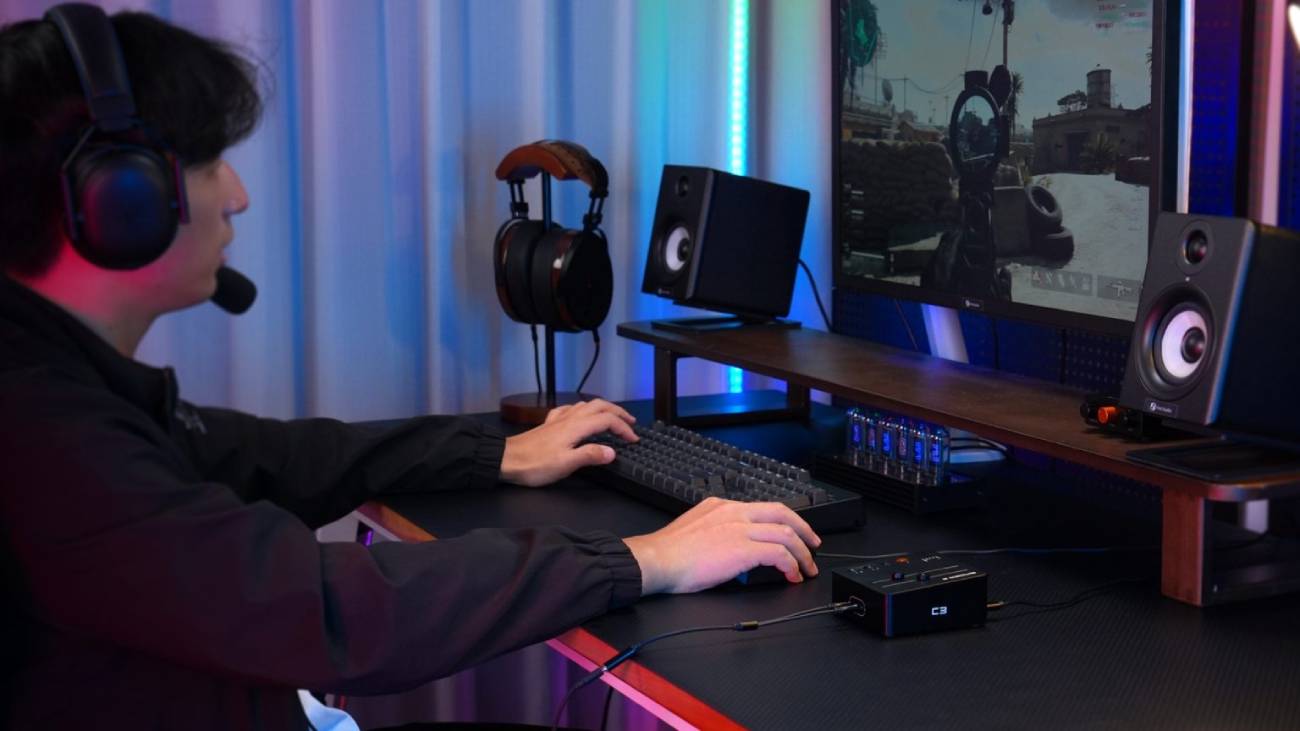 Joueur portant un casque-micro devant un écran de jeu FPS, la carte son gaming Fosi Audio C3 posée sur le bureau avec un casque branché en façade, entourée d'un clavier mécanique, d'enceintes de monitoring et d'un éclairage d'ambiance bleu et rouge