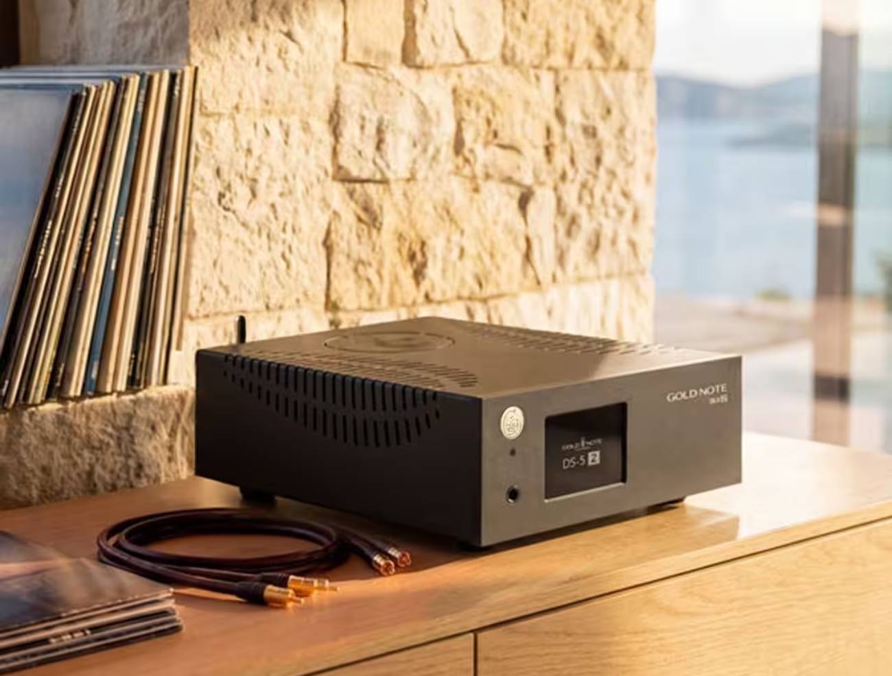 Streamer DAC Gold Note DS-5.2 en finition noire posé de trois-quarts sur un meuble en bois clair devant un mur en pierre, avec une collection de vinyles à gauche et un câble RCA à fiches cuivrées au premier plan, vue sur la mer en arrière-plan