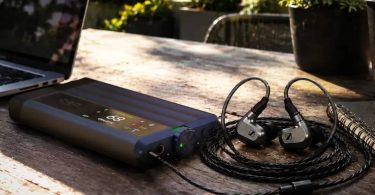 DAC et amplificateur casque portable iFi iDSD GR 2 en finition anthracite, écran OLED couleur affichant le volume, posé sur une table en bois à côté d'un ordinateur portable et d'écouteurs intra-auriculaires reliés en filaire.