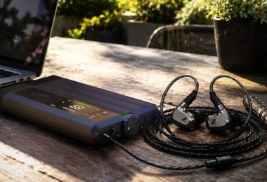 DAC et amplificateur casque portable iFi iDSD GR 2 en finition anthracite, écran OLED couleur affichant le volume, posé sur une table en bois à côté d'un ordinateur portable et d'écouteurs intra-auriculaires reliés en filaire.