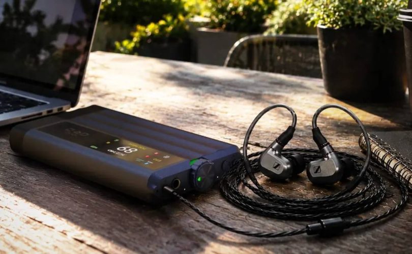 DAC et amplificateur casque portable iFi iDSD GR 2 en finition anthracite, écran OLED couleur affichant le volume, posé sur une table en bois à côté d'un ordinateur portable et d'écouteurs intra-auriculaires reliés en filaire.