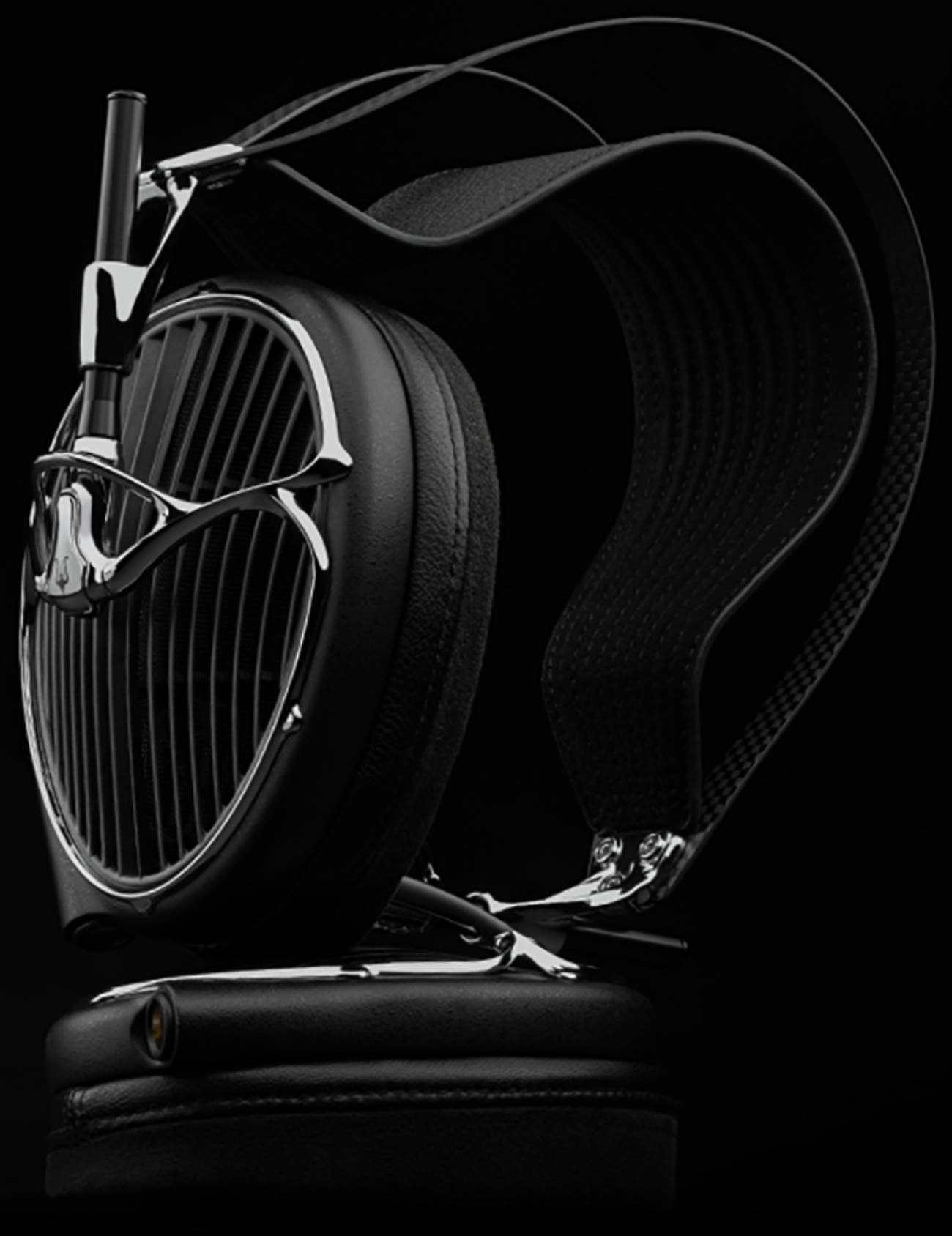 Vue de profil du casque Meze Audio ARTA noir posé sur un socle en cuir, montrant l'arceau suspendu en fibre de carbone avec sa sangle ajourée, la coque ouverte à grille et lames acoustiques, le cadre chromé et l'épais coussinet en cuir