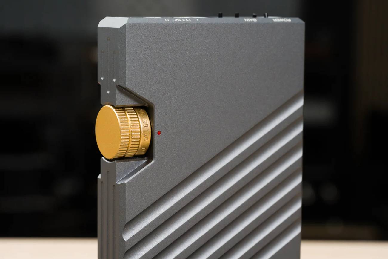 Gros plan sur l'amplificateur Oriolus DPA10 du système Traillii Ultra : molette de volume dorée graduée avec index rouge, châssis en aluminium gris aux rainures diagonales, et tranche supérieure portant les commandes POWER, GAIN et les sorties PHONE gauche et droite.