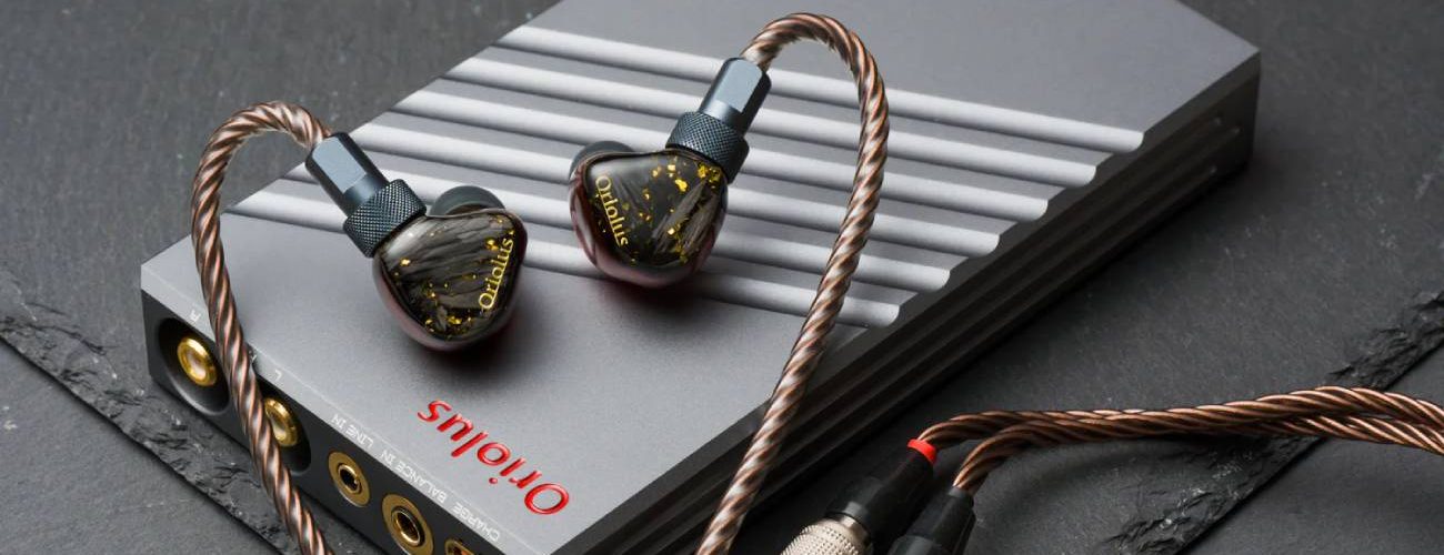 Système Oriolus Traillii Ultra : intra-auriculaires à coques sombres pailletées d'or et logo doré, posés sur l'amplificateur dédié DPA10 en aluminium gris rainuré à molette dorée, câble cuivré et connecteurs Hirose, sur un plan de marbre vert