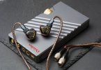 Système Oriolus Traillii Ultra : intra-auriculaires à coques sombres pailletées d'or et logo doré, posés sur l'amplificateur dédié DPA10 en aluminium gris rainuré à molette dorée, câble cuivré et connecteurs Hirose, sur un plan de marbre vert