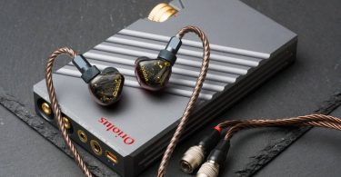Système Oriolus Traillii Ultra : intra-auriculaires à coques sombres pailletées d'or et logo doré, posés sur l'amplificateur dédié DPA10 en aluminium gris rainuré à molette dorée, câble cuivré et connecteurs Hirose, sur un plan de marbre vert
