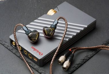 Système Oriolus Traillii Ultra : intra-auriculaires à coques sombres pailletées d'or et logo doré, posés sur l'amplificateur dédié DPA10 en aluminium gris rainuré à molette dorée, câble cuivré et connecteurs Hirose, sur un plan de marbre vert