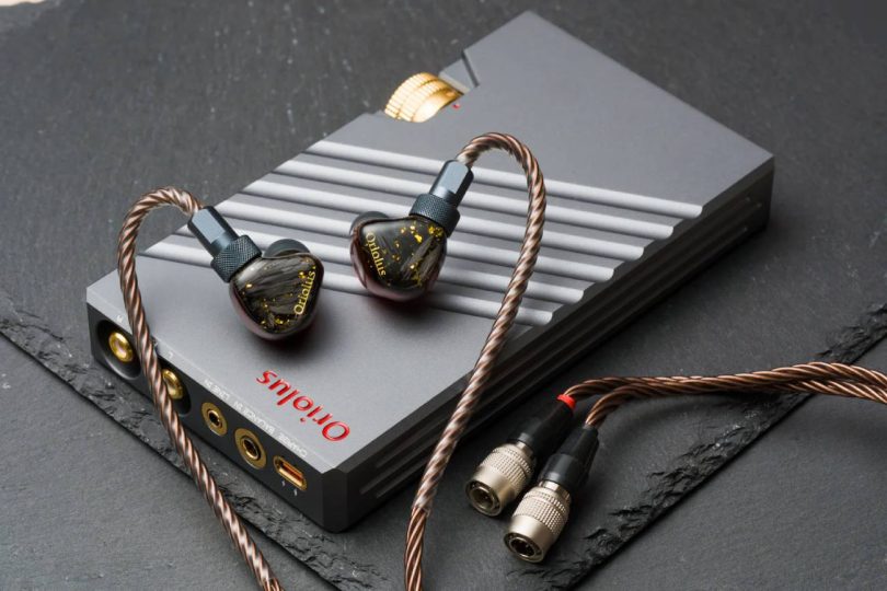 Système Oriolus Traillii Ultra : intra-auriculaires à coques sombres pailletées d'or et logo doré, posés sur l'amplificateur dédié DPA10 en aluminium gris rainuré à molette dorée, câble cuivré et connecteurs Hirose, sur un plan de marbre vert
