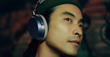 Homme de profil portant un casque circum-aural Sennheiser Momentum 5 Wireless, oreillette ovale aux accents métalliques argentés et coussinet sombre, dans une ambiance tamisée aux reflets verts et rouges, illustrant le confort d'écoute nomade premium