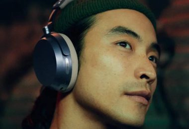 Homme de profil portant un casque circum-aural Sennheiser Momentum 5 Wireless, oreillette ovale aux accents métalliques argentés et coussinet sombre, dans une ambiance tamisée aux reflets verts et rouges, illustrant le confort d'écoute nomade premium
