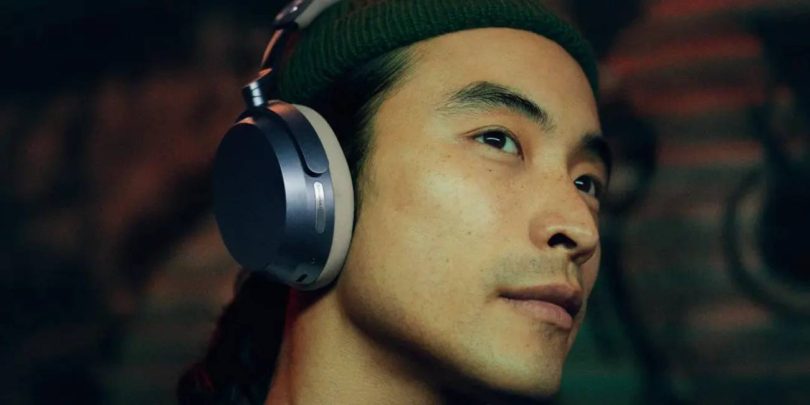 Homme de profil portant un casque circum-aural Sennheiser Momentum 5 Wireless, oreillette ovale aux accents métalliques argentés et coussinet sombre, dans une ambiance tamisée aux reflets verts et rouges, illustrant le confort d'écoute nomade premium
