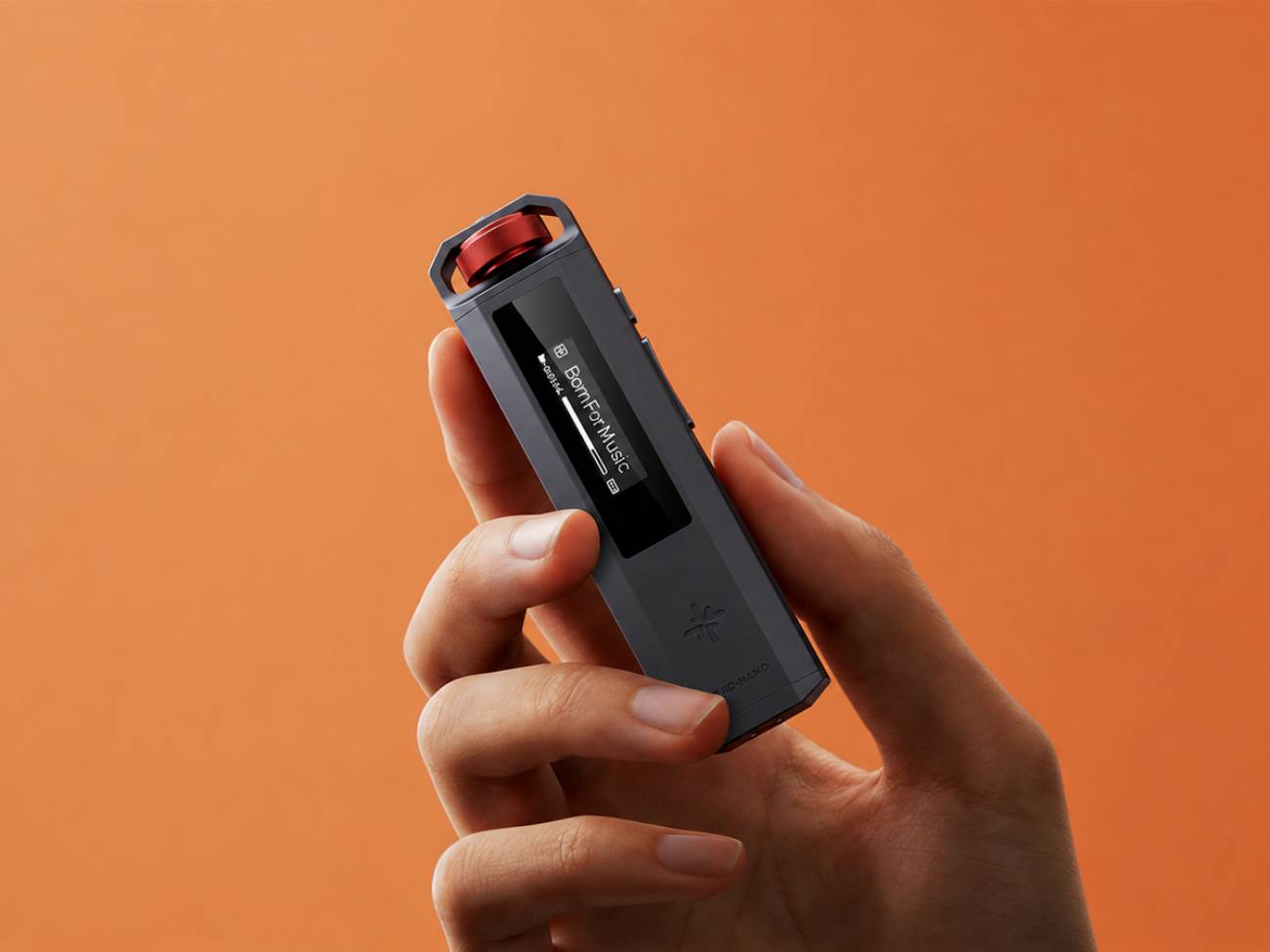Baladeur Snowsky ECHO NANO en finition noire tenu dans une main, molette multifonction rouge anodisée et boucle de lanière sur le dessus, écran OLED affichant Born For Music, boutons latéraux de lecture, sur fond orange