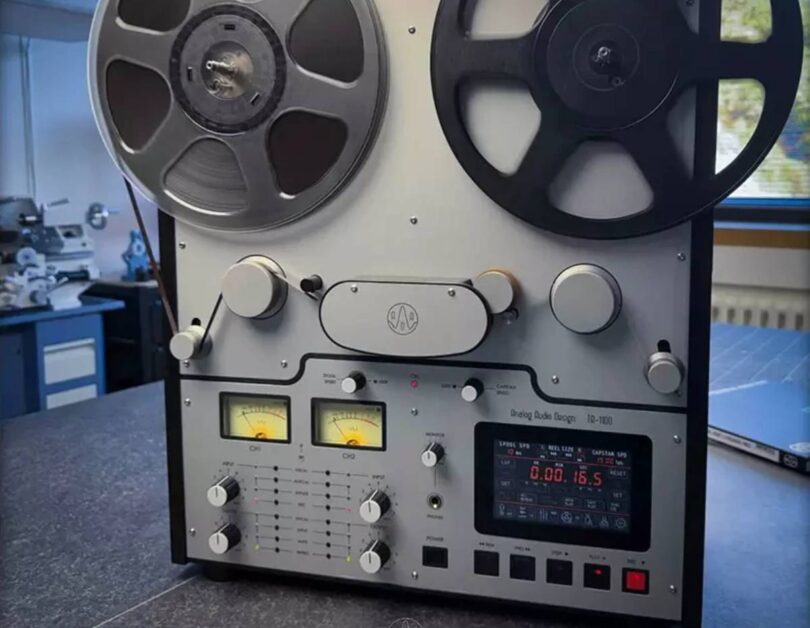 Magnétophone à bandes Analog Audio Design TR-1100 vu de face en atelier, bobine métal et bobine noire de 30 cm montées, deux vumètres à aiguille rétroéclairés jaune, afficheur numérique rouge, sélecteurs de vitesse bobines et cabestan, boutons de transport et touche REC rouge