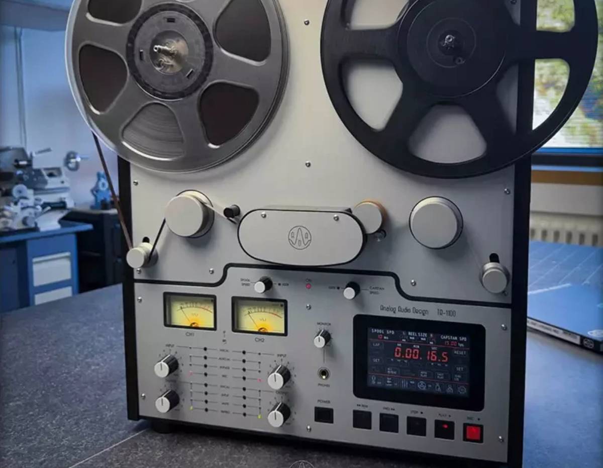 Magnétophone à bandes Analog Audio Design TR-1100 vu de face en atelier, bobine métal et bobine noire de 30 cm montées, deux vumètres à aiguille rétroéclairés jaune, afficheur numérique rouge, sélecteurs de vitesse bobines et cabestan, boutons de transport et touche REC rouge