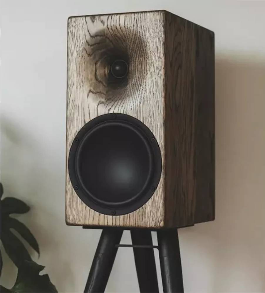 Enceinte bibliothèque Buchardt Audio E50-S en finition chêne fumé vue de trois quarts sur un pied noir à trois branches, montrant le tweeter dôme textile encastré dans son guide d'ondes creusé dans le baffle massif et le woofer Satori TeXtreme à cône noir