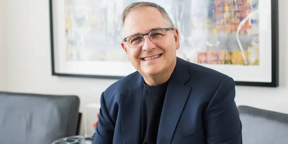 Portrait officiel de Marc Whitten, nouveau président et CEO de Dolby Laboratories, souriant en veste bleu marine et col roulé noir, assis dans un salon contemporain devant une toile abstraite colorée encadrée de noir