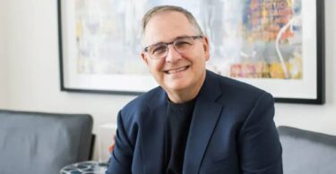 Portrait officiel de Marc Whitten, nouveau président et CEO de Dolby Laboratories, souriant en veste bleu marine et col roulé noir, assis dans un salon contemporain devant une toile abstraite colorée encadrée de noir