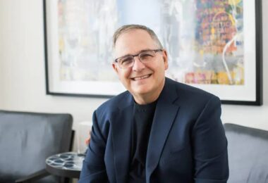 Portrait officiel de Marc Whitten, nouveau président et CEO de Dolby Laboratories, souriant en veste bleu marine et col roulé noir, assis dans un salon contemporain devant une toile abstraite colorée encadrée de noir