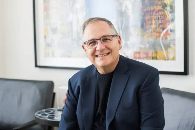 Portrait officiel de Marc Whitten, nouveau président et CEO de Dolby Laboratories, souriant en veste bleu marine et col roulé noir, assis dans un salon contemporain devant une toile abstraite colorée encadrée de noir