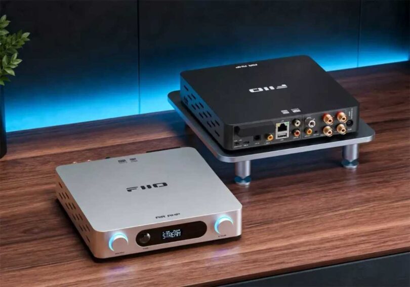 Deux amplificateurs de streaming FiiO Air Amp sur un bureau en bois : au premier plan la version argent vue de face avec son écran LCD affichant le mode STREAM et ses deux molettes rétroéclairées volume et treble, en arrière-plan la version noire vue de dos montrant le port Ethernet, les entrées USB, coaxiale et optique, les RCA Line In et le bornier haut-parleurs