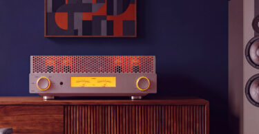 Amplificateur à tubes FiiO EA13 en finition silver posé sur un buffet en bois, capot perforé laissant voir les tubes EL34 incandescents, double vumètre mécanique rétroéclairé ambre et molette de volume à cerclage lumineux
