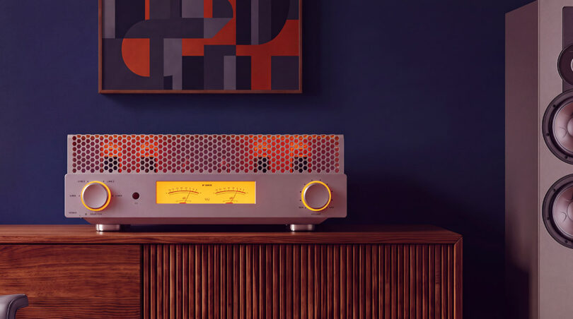 Amplificateur à tubes FiiO EA13 en finition silver posé sur un buffet en bois, capot perforé laissant voir les tubes EL34 incandescents, double vumètre mécanique rétroéclairé ambre et molette de volume à cerclage lumineux