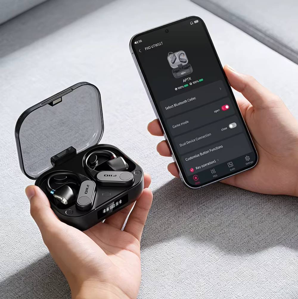 Boîtier de charge ouvert du FiiO UTWS17 tenu dans une main avec les deux contours d'oreille noirs à l'intérieur, face à un smartphone affichant l'application FiiO Control : codec aptX actif, niveaux de batterie à 100 pour cent, sélection du codec Bluetooth, mode jeu, double connexion et personnalisation des boutons