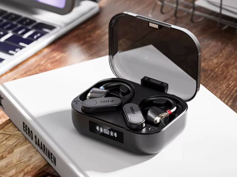 Paire de contours d'oreille Bluetooth FiiO UTWS17 en finition noire posée dans son boîtier de charge à couvercle transparent fumé, écran OLED affichant les niveaux de batterie et l'état d'appairage, connecteurs MMCX rouge et bleu visibles sur les embouts