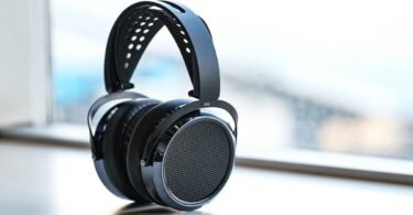 Casque HIFIMAN HE6 Remastered planaire magnétique ouvert en finition noire posé sur fond blanc, vue trois quarts montrant la grille circulaire ajourée du transducteur, le contour laqué anthracite, le bandeau suspendu perforé en suédine et l'arceau composite marqué HE6