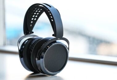 Casque HIFIMAN HE6 Remastered planaire magnétique ouvert en finition noire posé sur fond blanc, vue trois quarts montrant la grille circulaire ajourée du transducteur, le contour laqué anthracite, le bandeau suspendu perforé en suédine et l'arceau composite marqué HE6