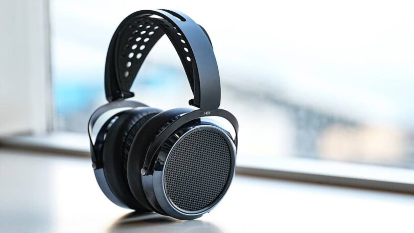 Casque HIFIMAN HE6 Remastered planaire magnétique ouvert en finition noire posé sur fond blanc, vue trois quarts montrant la grille circulaire ajourée du transducteur, le contour laqué anthracite, le bandeau suspendu perforé en suédine et l'arceau composite marqué HE6