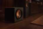 Caisson de basses compact Klipsch RP-1600CSW Reference Premiere à charge close posé au sol près d'une colonne Klipsch, woofer Cerametallic cuivré frontal sans évent, finition vinyle ébène