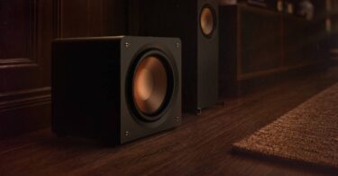 Caisson de basses compact Klipsch RP-1600CSW Reference Premiere à charge close posé au sol près d'une colonne Klipsch, woofer Cerametallic cuivré frontal sans évent, finition vinyle ébène
