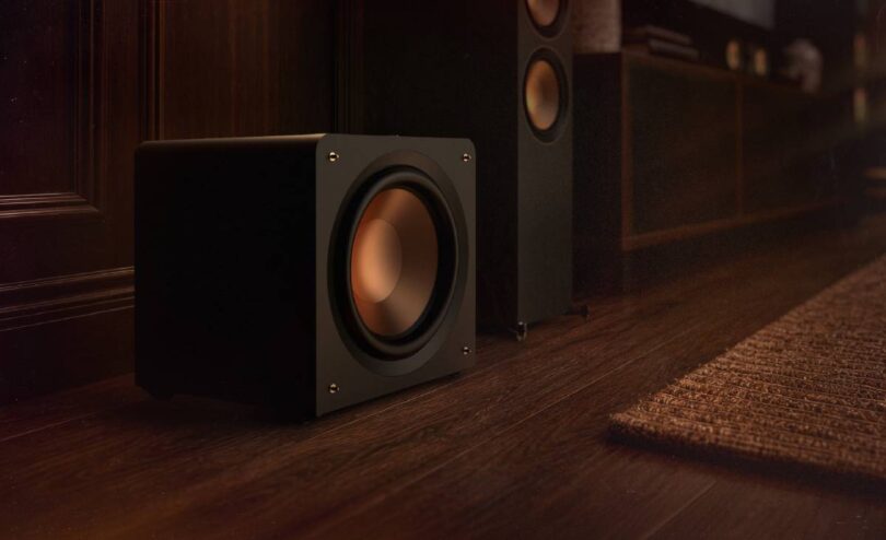 Caisson de basses compact Klipsch RP-1600CSW Reference Premiere à charge close posé au sol près d'une colonne Klipsch, woofer Cerametallic cuivré frontal sans évent, finition vinyle ébène