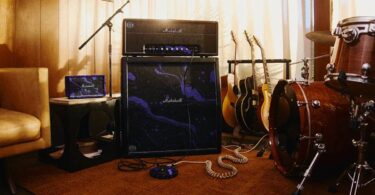 Tête d'ampli Marshall JMH Studio Hendrix 60th Anniversary posée sur un baffle 4x12 recouvert du motif psychédélique violet oil-on-water, avec pédalier assorti au sol, enceinte portable Marshall violette sur une table d'appoint, guitares acoustiques et batterie dans un studio de répétition