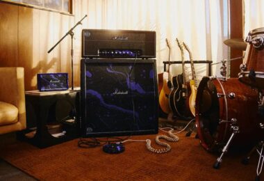Tête d'ampli Marshall JMH Studio Hendrix 60th Anniversary posée sur un baffle 4x12 recouvert du motif psychédélique violet oil-on-water, avec pédalier assorti au sol, enceinte portable Marshall violette sur une table d'appoint, guitares acoustiques et batterie dans un studio de répétition