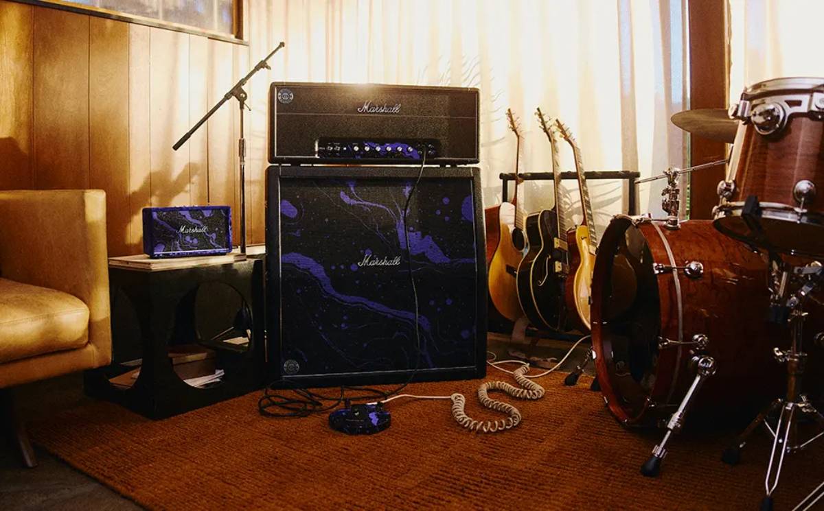 Tête d'ampli Marshall JMH Studio Hendrix 60th Anniversary posée sur un baffle 4x12 recouvert du motif psychédélique violet oil-on-water, avec pédalier assorti au sol, enceinte portable Marshall violette sur une table d'appoint, guitares acoustiques et batterie dans un studio de répétition