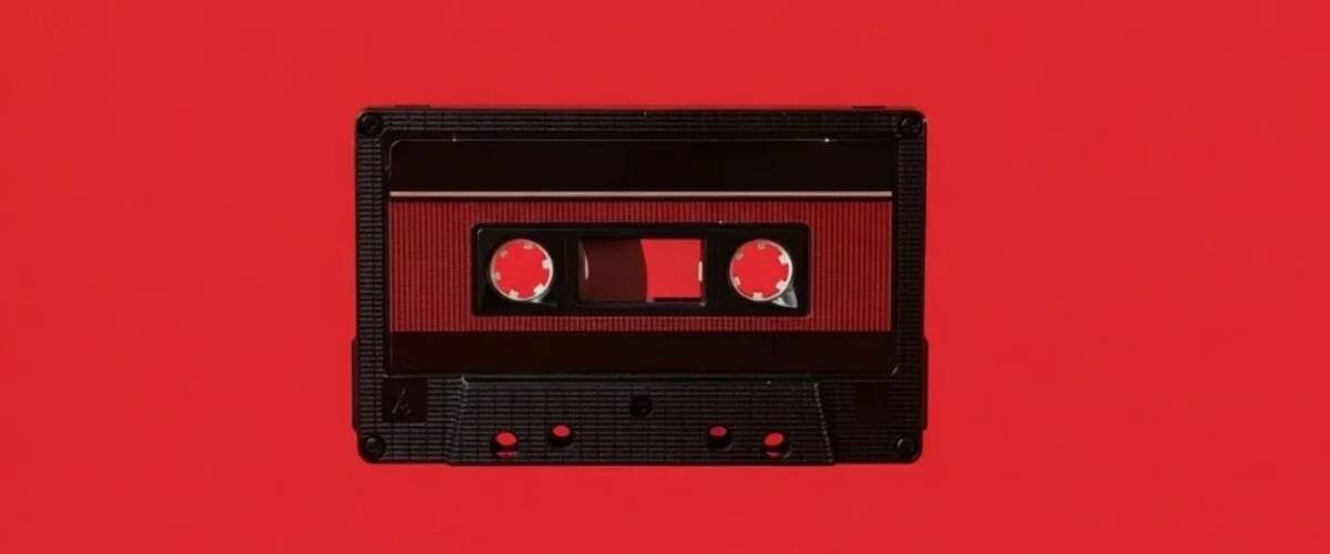 Cassette audio Maxell UD-60U en édition limitée Disk Union photographiée à plat sur fond rouge vif, coquille noire à texture nervurée, fenêtre centrale laissant voir la bande ferrique et les deux moyeux dentés, étiquette rouge sombre et repères de vis apparents