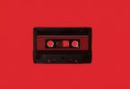 Cassette audio Maxell UD-60U en édition limitée Disk Union photographiée à plat sur fond rouge vif, coquille noire à texture nervurée, fenêtre centrale laissant voir la bande ferrique et les deux moyeux dentés, étiquette rouge sombre et repères de vis apparents