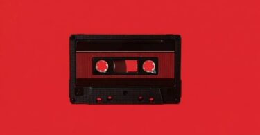 Cassette audio Maxell UD-60U en édition limitée Disk Union photographiée à plat sur fond rouge vif, coquille noire à texture nervurée, fenêtre centrale laissant voir la bande ferrique et les deux moyeux dentés, étiquette rouge sombre et repères de vis apparents