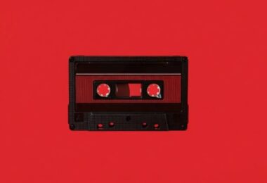 Cassette audio Maxell UD-60U en édition limitée Disk Union photographiée à plat sur fond rouge vif, coquille noire à texture nervurée, fenêtre centrale laissant voir la bande ferrique et les deux moyeux dentés, étiquette rouge sombre et repères de vis apparents