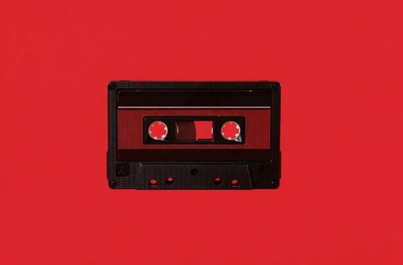 Cassette audio Maxell UD-60U en édition limitée Disk Union photographiée à plat sur fond rouge vif, coquille noire à texture nervurée, fenêtre centrale laissant voir la bande ferrique et les deux moyeux dentés, étiquette rouge sombre et repères de vis apparents