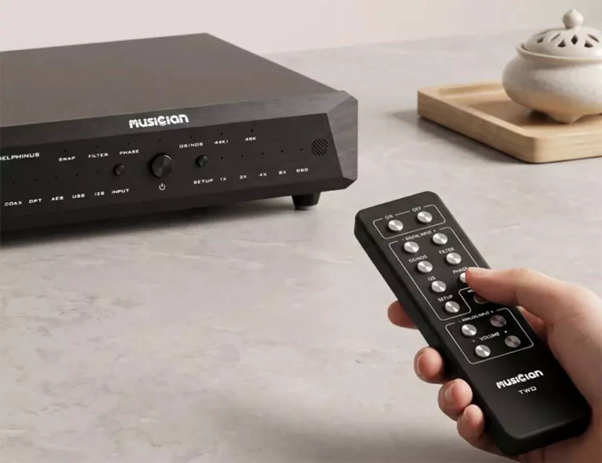 Main tenant la télécommande Musician TWO devant le convertisseur R-2R Delphinus noir, avec les touches ON et OFF en haut, le bloc DIGITAL INPUT regroupant OS/NOS, FILTER, I2S, PHASE et SETUP, et le bloc ANALOG INPUT avec les commandes de volume, la façade du DAC affichant les indicateurs SWAP, FILTER, PHASE, OS/NOS et les fréquences 44K1 et 48K