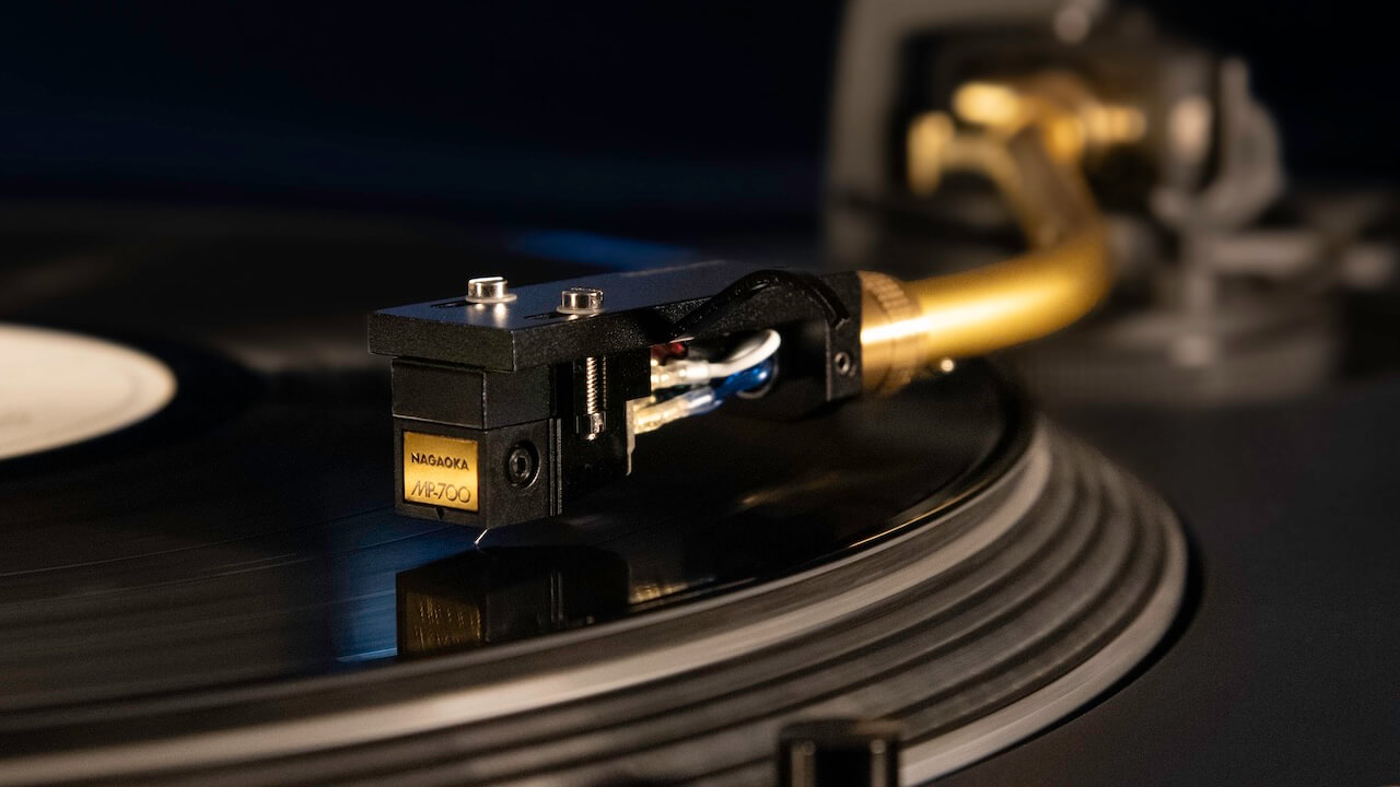 Cellule phono Nagaoka MP-700 montée sur un porte-cellule doré au bout du bras de lecture d'une platine vinyle, diamant nu MicroRidge en contact avec le sillon du disque en rotation, fils de liaison rouge, bleu, blanc et vert visibles sur les broches