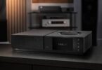 Amplificateur streaming Naim Uniti Star Lite en finition noire vu de trois quarts sur un meuble sombre, avec écran couleur 5 pouces affichant le logo Starlite, molette de volume circulaire sur le capot, port USB frontal, quatre touches tactiles et ailettes de dissipation latérales