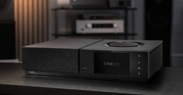 Amplificateur streaming Naim Uniti Star Lite en finition noire vu de trois quarts sur un meuble sombre, avec écran couleur 5 pouces affichant le logo Starlite, molette de volume circulaire sur le capot, port USB frontal, quatre touches tactiles et ailettes de dissipation latérales