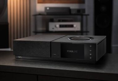 Amplificateur streaming Naim Uniti Star Lite en finition noire vu de trois quarts sur un meuble sombre, avec écran couleur 5 pouces affichant le logo Starlite, molette de volume circulaire sur le capot, port USB frontal, quatre touches tactiles et ailettes de dissipation latérales