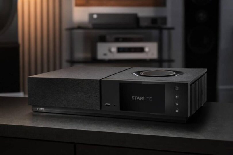 Amplificateur streaming Naim Uniti Star Lite en finition noire vu de trois quarts sur un meuble sombre, avec écran couleur 5 pouces affichant le logo Starlite, molette de volume circulaire sur le capot, port USB frontal, quatre touches tactiles et ailettes de dissipation latérales
