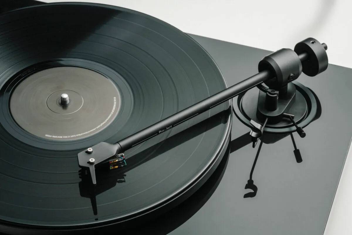 Gros plan sur le bras aluminium 8,6 pouces de la Pro-Ject E1.2 en finition High Gloss Black avec un disque vinyle en place, montrant le porte-cellule et la cellule Pick it MM E, le cardan, le contrepoids arrière et l'antiskating à fil et poids suspendu